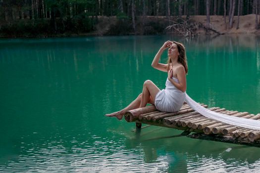 A girl with fluffy hair in a white light dress barefoot in the summer in a forest on a lake sits on a pantone on a wooden pier bridge. The concept of harmony with nature, leisure and travel, blogger
