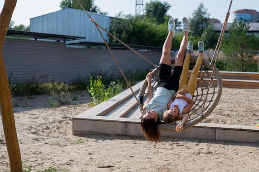 Mom and daughter swing on a round swing. Caucasian woman and little girl have fun on the playground