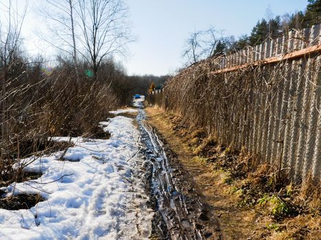 Abandoned dirty village street with snow in spring.View of a village small street with a fence.