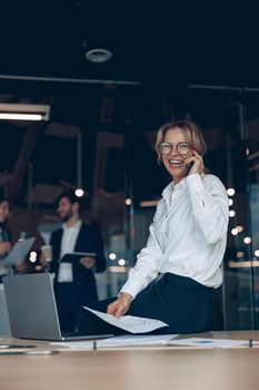 mature business woman talking on phone during meeting in office with colleagues in the background