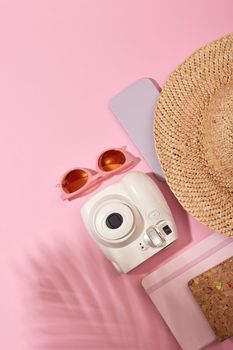 From above of instant photo camera with sunglasses and straw hat placed near notebook on pink background with shadow of plant