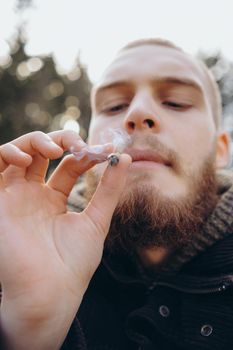 Young man beard smokes cigarette or tobacco. Guy is addicted to cigarettes and always smokes lot of tobacco every day. He smokes cigarette outdoors on background of lake in park. Cool guy blows smoke