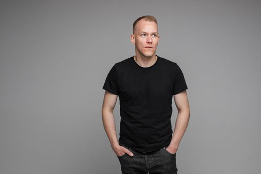 picture of caucasian man keeps his hands in the pockets of his black jeans and looks to the right isolated on grey background