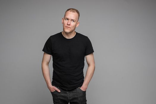 picture of caucasian man keeps his hands in the pockets of his black jeans and looks to the right isolated on grey background