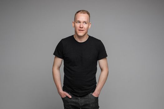 picture of caucasian man keeps his hands in the pockets of his black jeans and looks to the right isolated on grey background