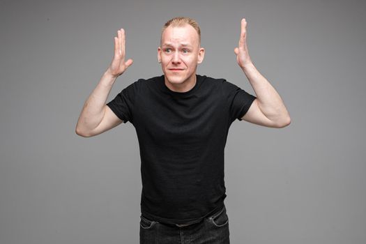 Portrait of puzzled blond Caucasian man in black t-shirt holding arms up showing misunderstanding and disbelief. He doesn’t understand what is happening.