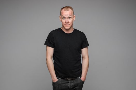 Portrait of a man in black looking at camera with amazed wide open eyes holding hands in pockets of his black jeans. Isolate on grey background.