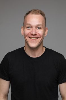 Studio portrait of adult man with short blond hair wearing casual black t-shirt smiling cheerfully at camera with friendly look. Cutout on grey background.