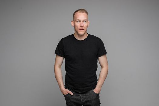 picture of caucasian man keeps his hands in the pockets of his black jeans and looks to the right isolated on grey background