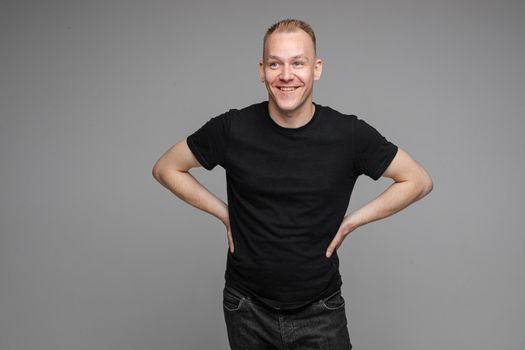 Portrait of blond Caucasian man in black t-shirt and dark jeans holding hands together with eyes closed on grey background. Bored or tired asleep man standing on grey background.