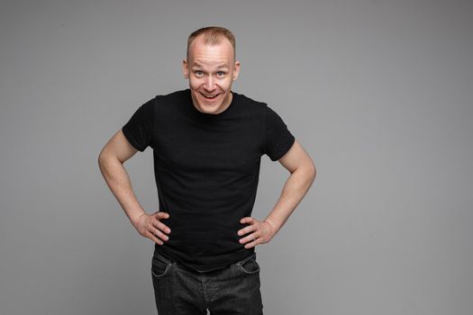 Surprised Caucasian man in black looking at camera with smile with hands on his waist.