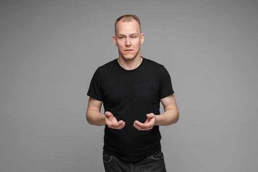 Portrait of blond Caucasian man in black t-shirt and dark jeans holding hands together with eyes closed on grey background. Bored or tired asleep man standing on grey background.