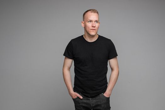 picture of caucasian man keeps his hands in the pockets of his black jeans and looks to the right isolated on grey background