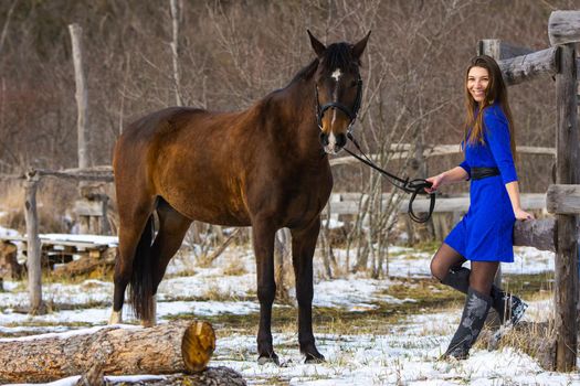 A beautiful girl in a short blue dress walks with a horse at an old farm in winter, the girl smiles and looks into the frame a