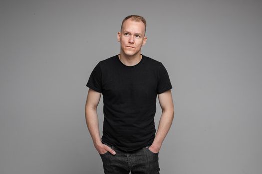 picture of caucasian man keeps his hands in the pockets of his black jeans and looks to the right isolated on grey background