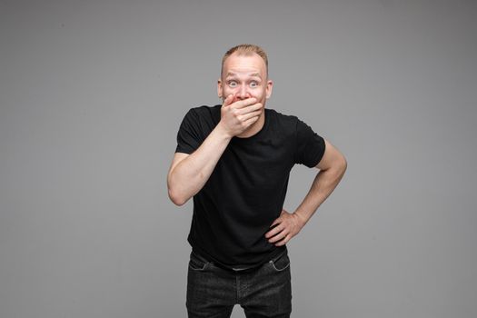 Studio portrait of excited blond Caucasian man in black t-shirt and jeans covering his mouth with hand in astonishment. Isolate on grey background.