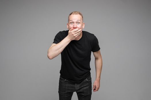 Studio portrait of excited blond Caucasian man in black t-shirt and jeans covering his mouth with hand in astonishment. Isolate on grey background.