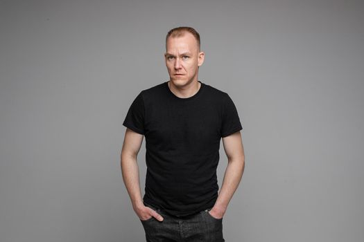 picture of caucasian man keeps his hands in the pockets of his black jeans and looks to the left isolated on grey background