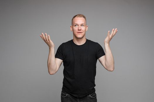 Studio portrait of excited blond Caucasian man in black t-shirt and jeans covering his mouth with hand in astonishment. Isolate on grey background.