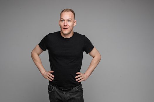 Surprised Caucasian man in black looking at camera with smile with hands on his waist.