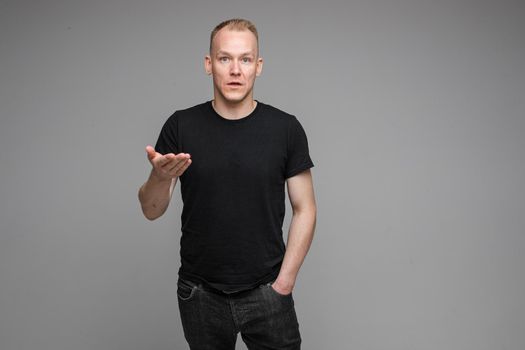 Portrait of puzzled and confused man in black t-shirt and jeans gesturing at camera with his hand showing misunderstanding.