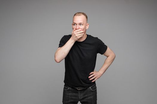 Studio portrait of excited blond Caucasian man in black t-shirt and jeans covering his mouth with hand in astonishment. Isolate on grey background.