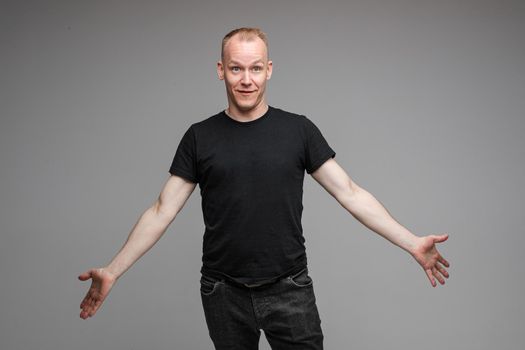 Studio portrait of handsome adult Caucasian man with short blond hair in black t-shirt and dark jeans looking away with puzzled face and outstretched arms...