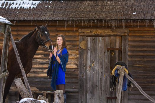 A girl stands with a horse against the background of an old wooden house a