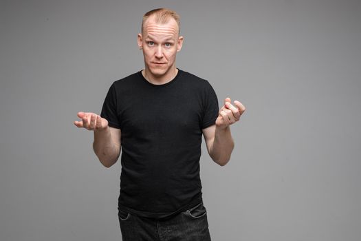 Portrait of Caucasian blond man in dark clothes outstretching his arms while showing his tongue at camera with wide open eyes. Cutout on grey background.