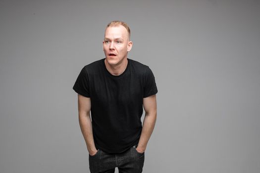 Studio portrait of handsome adult Caucasian man with short blond hair in black t-shirt and dark jeans looking away with puzzled face and outstretched arms.