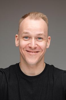 Close-up photo of friendly fair-haired man smiling while posing for a camera at a studio