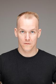 Studio portrait of adult blond Caucasian man with short haircut in black t-shirt with blue eyes staring at camera on grey background.