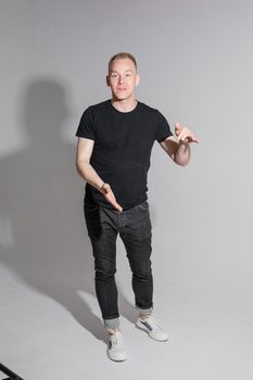 Full length of adult Caucasian blonde man in black t-shirt, dark jeans and sneakers talking to camera with arms bent, holding fists. Presenter or advertiser promoting something.