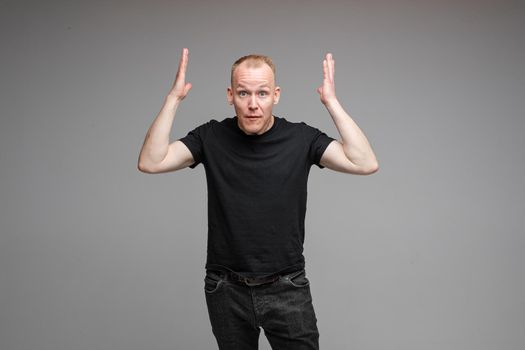 Studio portrait of handsome adult Caucasian man with short blond hair in black t-shirt and dark jeans looking away with puzzled face and outstretched arms.