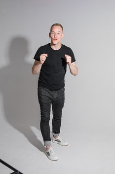 Full length of adult Caucasian blonde man in black t-shirt, dark jeans and sneakers talking to camera with arms bent, holding fists. Presenter or advertiser promoting something.