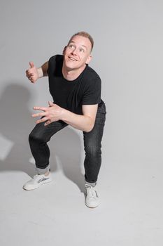 Full length of adult Caucasian blonde man in black t-shirt, dark jeans and sneakers talking to camera with arms bent, holding fists. Presenter or advertiser promoting something.