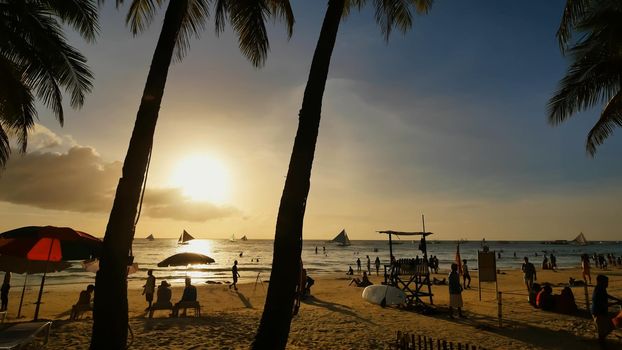 Beach with silhouettes of tourists among palm trees on the island of Boracay. Palm trees in the rays of sunset. Sailboats on the water. Philippine Tropics. Shooting in motion