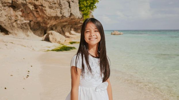 A charming and happy philippine teenage girl in a white summer dress is running along a tropical beach near the rocks. She is happily spinning. Childhood. Recreation