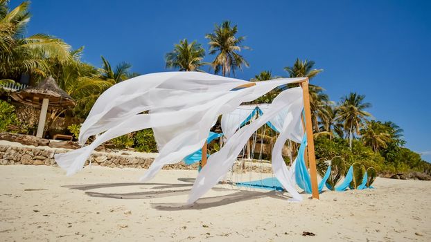 Wedding arch decorated with flowers on the beach near the ocean. Her white and blue canvases fly in the wind. Decorative hearts from the leaves of palm trees. Sandy tropical shore