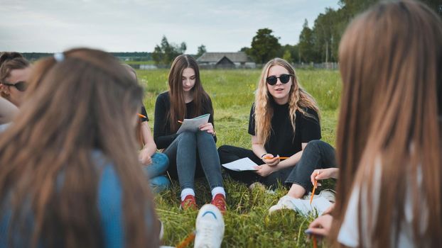 A group of female students are sitting in a circle on a meadow for collective work with notebooks