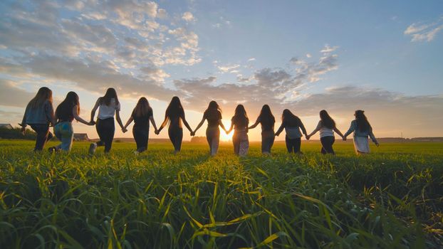 A group of girls walk towards the sun at sunset holding hands