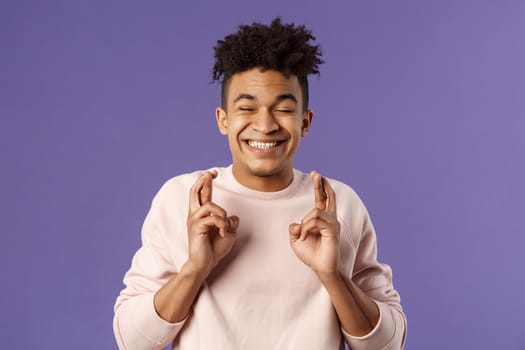 Close-up portrait of happy young hopeful guy anticipating something good happen, cross fingers good luck, close eyes and smiling awaiting miracle, praying for dream come true, purple background.