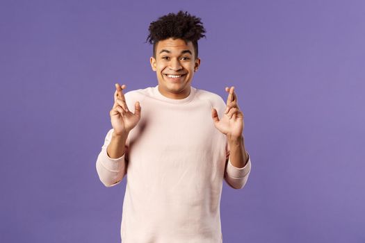 Portrait of hopeful positive, smiling young man having faith in dreams may come true, make wish, cross fingers good luck and awaiting for miracle, standing purple background praying.