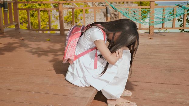 An philippine schoolgirl girl with a backpack is sitting and crying near the tropical coast. Sad mood