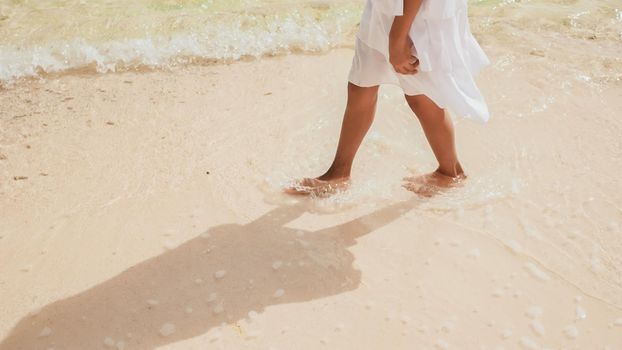 The tanned legs of an philippine schoolgirl in a white dress run along white sand, touching the waves of foam on the beach. Tropical landscape. Childhood