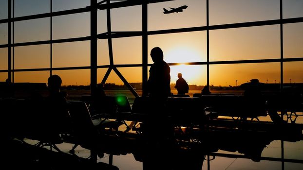 Silhouette of a tourist guy watching the take-off of the plane standing at the airport window at sunset in the evening. Travel concept, people in the airport