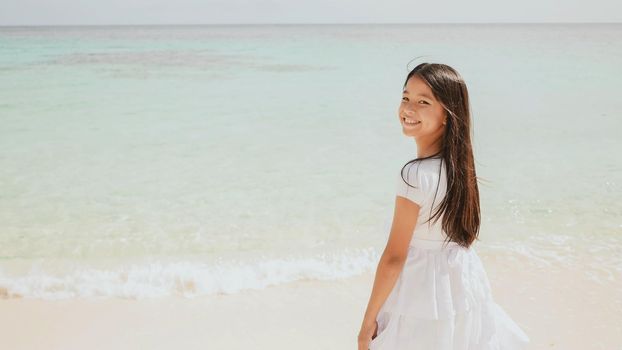 A charming philippine schoolgirl in a white dress is walking along a white sandy beach. Enjoying the tropical scenery. Childhood