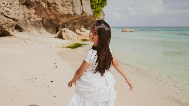 A charming and happy philippine teenage girl in a white summer dress is running along a tropical beach near the rocks. She is happily spinning. Childhood. Recreation