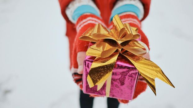 Young charming schoolgirl in mittens gives a gift, looking at the camera in the winter forest. A wrapped box with a gift in the hands of a schoolgirl close-up