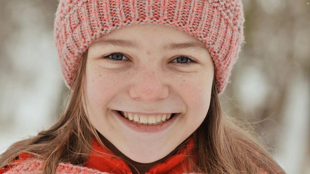Portrait of a young schoolgirl with freckles in the woods in winter. The girl touches her nose with her hand in the mitten